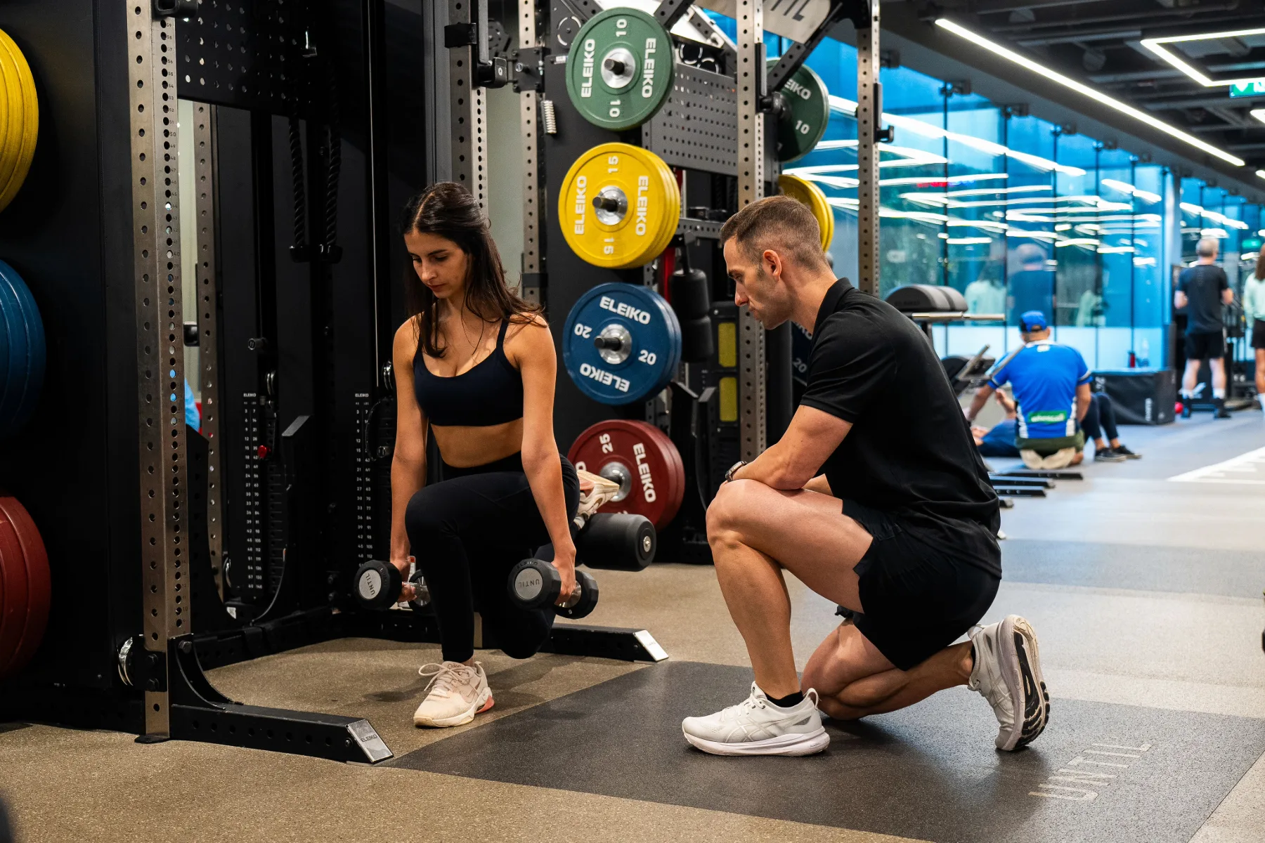 Maurizio crouching beside a client performing a dumbbell squat in the gym